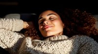 Young Hispanic woman with curly hair relaxing in cozy knit sweater, eyes closed with peaceful expression, bathed in warm natural sunlight. Perfect for wellness content.