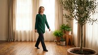 Mature woman in green blazer walking through bright living room with potted plants and natural light from windows.