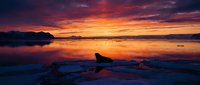 Dramatic sunset over calm lake with perfect reflections creating mirror effect on water surface. Silhouetted rocks and distant mountains under colorful sky.