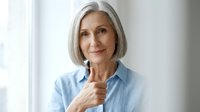 Mature woman with gray hair wearing light blue shirt, smiling confidently at camera in bright studio setting. Portrait for healthcare, wellness, business and lifestyle content.