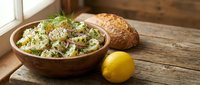 Wooden bowl filled with fresh green salad, whole grain bread roll, and lemon on rustic wooden table near window for food styling and culinary content.