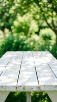 Empty white wooden table with blurred green foliage background, suitable for product placement, outdoor advertising, and garden-themed designs.