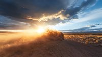 Dramatic desert landscape at sunset with sunburst through storm clouds, golden light illuminating dusty terrain and rocky outcrop in wilderness setting.