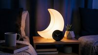 Modern crescent moon night light glowing on wooden bedside table in cozy bedroom interior with clock and plant creating ambient atmosphere for relaxation.