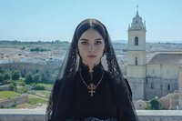 Young woman in traditional black lace mantilla veil with ornate cross necklace standing against historic Mediterranean cityscape with bell tower and ancient architecture.