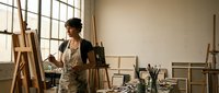 Young Asian woman artist painting on canvas in bright art studio with easels, brushes and creative supplies for artistic projects.