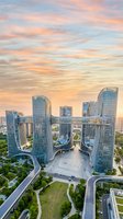 Aerial view of modern curved glass skyscrapers with central plaza at sunset, surrounded by green spaces and connecting roads, ideal for corporate and urban development projects.