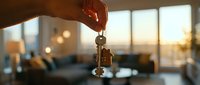 Hand holding house keys with a home-shaped keychain in a modern apartment interior at golden sunset, symbolizing real estate purchase or rental.