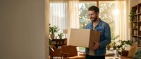 Young Caucasian man in denim shirt holding cardboard box while moving into new apartment with warm sunlight streaming through windows.