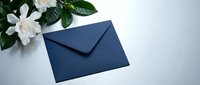 Navy blue envelope on a light gray surface beside white gardenia flowers, elegant stationery concept for wedding invitations or sympathy cards.