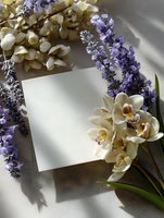 Blank white card with fresh orchids and lavender flowers on light background with natural shadows, elegant floral composition for wedding invitations.