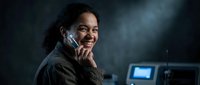 Young African American woman smiling while talking on smartphone in modern office environment with computer screens and atmospheric lighting.