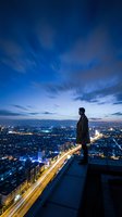 Man standing on rooftop overlooking illuminated cityscape at dusk, wearing dark coat, gazing at urban skyline with glowing streets and buildings below during twilight hour.