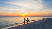 Romantic couple walking hand in hand on pristine beach during golden sunset with warm ocean waves and colorful sky for vacation and travel concepts.