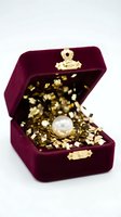 Burgundy velvet jewelry box with gold-toned metal clasp, open to reveal pearl gemstone nestled in gold foil confetti, isolated on white background for jewelry retail and luxury product design.