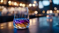 Colorful cocktail in modern twisted glass with vibrant blue and purple reflections against blurred bokeh lights background on dark wooden bar counter.