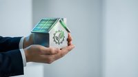 Businessman hands holding miniature eco house model with solar panels and green plant symbol representing sustainable energy solutions.