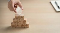 Hand placing wooden block with house icon on pyramid of insurance symbol cubes representing property protection and risk management services.