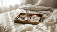Cozy morning bedroom scene with wooden breakfast tray on unmade bed featuring coffee cup, dried flowers and good morning love note card in soft natural light.