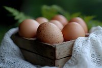 Fresh brown eggs in rustic wooden box with white linen cloth and green leaves, shallow depth of field creates soft dreamy mood for farm fresh concept.