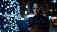 Young African American woman with curly hair and glasses using smartphone at night with bokeh city lights background for technology and communication concepts.