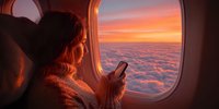 Young woman in cozy sweater using smartphone on airplane, gazing at stunning sunset over clouds through window.