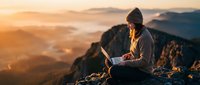 Person sitting on mountain peak using laptop at sunrise with misty valley below, remote work and digital nomad lifestyle concept for business and technology themes.
