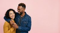 Young diverse couple embracing against pink background. Woman with dark curly hair wearing yellow sweater, man with beard in denim shirt. Studio portrait for relationship, love, and lifestyle advertising.