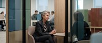 Senior woman with gray hair, wearing black blazer, engaged in professional conversation with female colleague in modern office meeting room with glass partitions and wooden accents. Suitable for corporate training, business communication, and workplace diversity content.