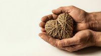 Hands gently holding woven twine heart symbol representing love, care, protection and emotional support in warm natural lighting.