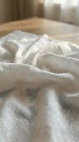 Soft white linen fabric with natural wrinkles and folds on wooden surface in warm morning light creating cozy textile background for home decor projects.