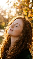 Young woman with curly red hair enjoying warm sunlight in autumn park with golden bokeh background. Peaceful moment of mindfulness and relaxation outdoors.