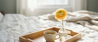 Morning coffee in bed with yellow ranunculus flower on wooden tray creating cozy bedroom atmosphere for relaxation and self-care lifestyle moments.