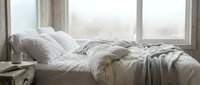 Unmade bed with white rumpled linens and pillows near large windows with natural daylight, suitable for home decor and lifestyle content.