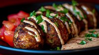 Juicy grilled meatballs drizzled with creamy sauce and fresh herbs served on dark plate with cherry tomatoes, close up food photography.