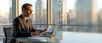 Professional businessman in suit working on laptop in modern office with city skyline view through windows during golden hour lighting.