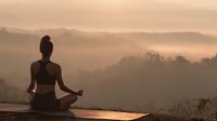 Young woman practicing yoga meditation in lotus pose at sunrise with misty mountain landscape background creating peaceful wellness atmosphere.