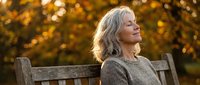 Peaceful mature woman with gray hair relaxing on wooden bench during golden hour autumn sunset, eyes closed in serene meditation outdoors.