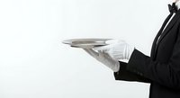 Professional waiter in black suit holding silver serving tray with white gloves against clean background for hospitality service concepts.