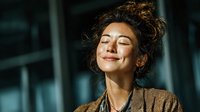 Young professional woman with curly hair smiling peacefully with eyes closed in modern office environment, expressing joy and workplace satisfaction.