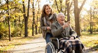Happy senior man in wheelchair with caring female caregiver enjoying autumn day in park. Elderly care, disability support, healthcare assistance concept.