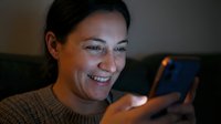 Middle-aged woman with dark hair and olive complexion, smiling while holding and looking at a smartphone in warm indoor lighting, suitable for technology, communication, and lifestyle content.