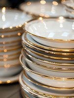 Stack of elegant white porcelain plates with gold trim edges against blurred bokeh lights background, creating luxurious dining atmosphere for events and celebrations.