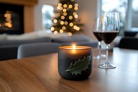Cozy winter evening with glowing holly leaf candle and red wine glass on wooden table, Christmas tree lights bokeh in background creating warm atmosphere.