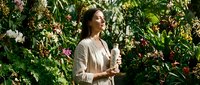Young professional woman in beige blazer holding coffee cup in lush tropical garden with flowering plants, enjoying peaceful morning break outdoors.