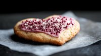 Heart shaped sugar cookie with pink frosting and red sprinkles on parchment paper against dark background. Romantic dessert with selective focus.