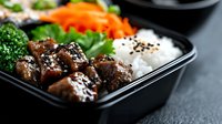 Glazed teriyaki chicken with white rice, steamed broccoli and carrots in black bento box. Asian cuisine meal prep with sesame seeds garnish on dark background.