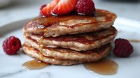 Stack of homemade fluffy pancakes topped with fresh red raspberries and drizzled with golden maple syrup on white plate, close up view of breakfast food.