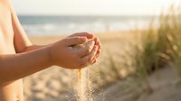 Woman hands cupping and releasing golden sand at sunny beach with ocean waves and dune grass in soft focus background during golden hour vacation moment.