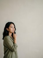 Thoughtful young woman with long black hair and East Asian ethnicity wearing olive green linen shirt, hand near face in contemplative pose against neutral beige wall background.